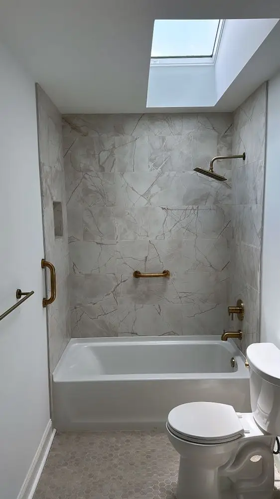 Bathroom with a tub under a skylight, marble-look tile surround and brushed brass fixtures — a completed CMD Property Restoration Group project