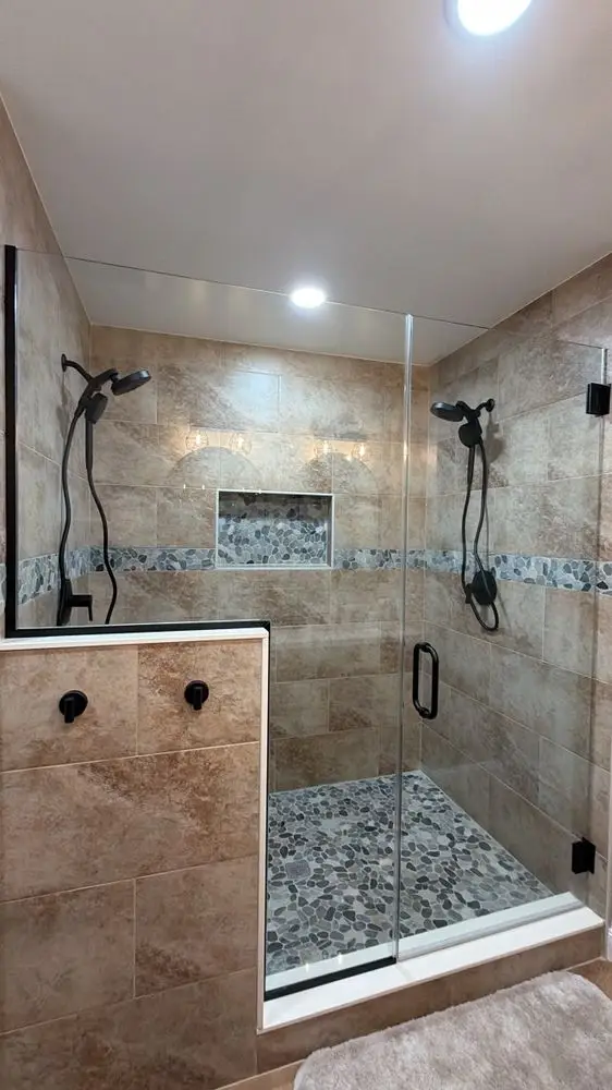 Walk-in shower with travertine-look tile, a glass mosaic accent band, dual shower heads and pebble-style floor — a completed CMD Property Restoration Group project