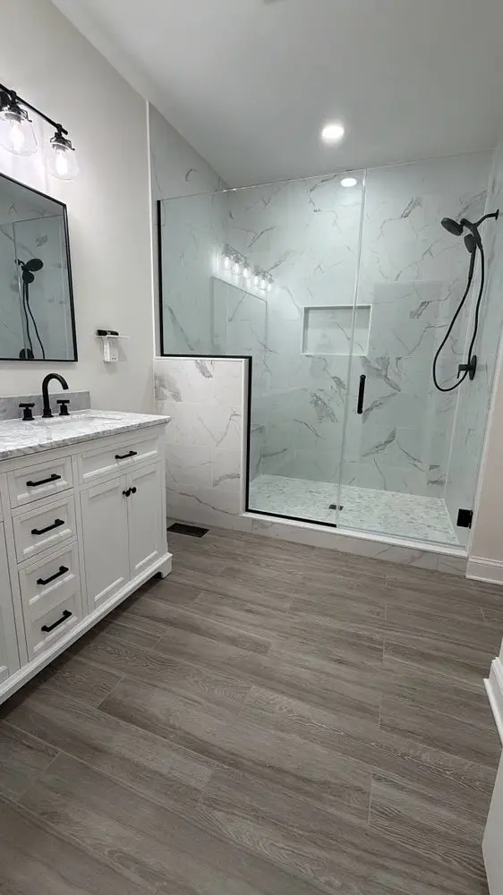Bathroom with a white vanity, frameless glass shower with marble-look tile, black fixtures and wood-look plank floor — a completed CMD Property Restoration Group project