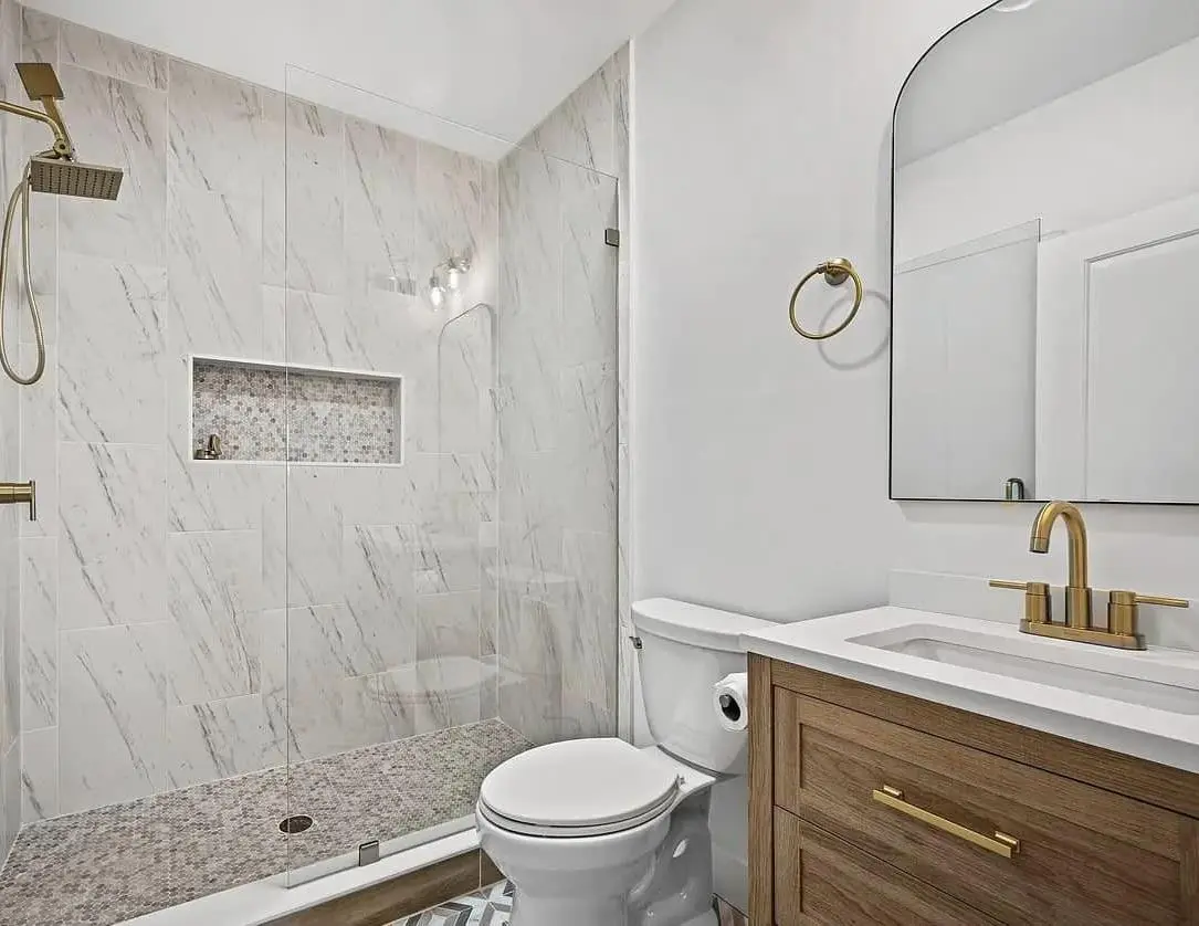 Bathroom with a marble-look tile shower, brass fixtures, oak vanity and arched mirror — a completed CMD whole-home renovation