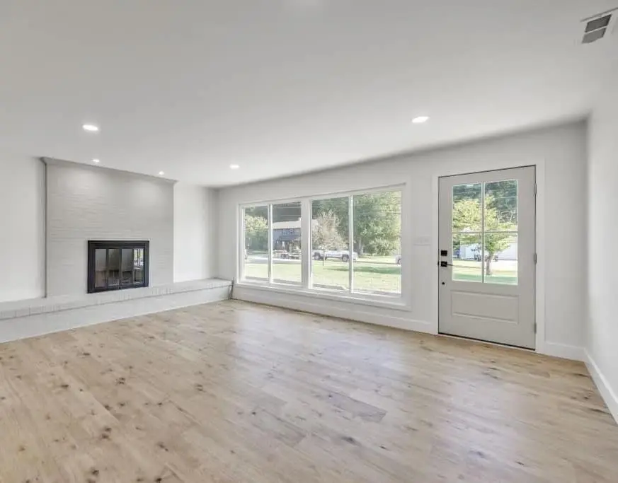 Living room with a painted brick fireplace, raised hearth, large windows and light oak plank floors — a completed CMD whole-home renovation